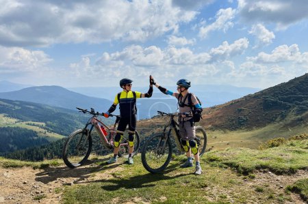 Two cyclists men riding electric bikes outdoors. Male tourists resting on the top of hill, giving high five to each other, enjoying beautiful mountain landscape, wearing helmet and backpack.