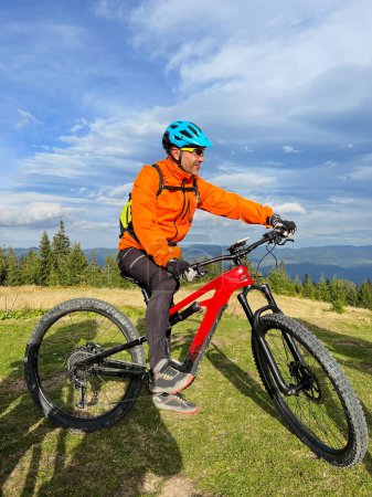Cyclist man riding electric bike outdoors on sunny day. Male tourist resting on grassy hill, enjoying beautiful mountain landscape, wearing helmet and backpack. Concept of active leisure.