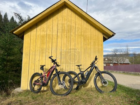 Two modern electric mountain bikes leaning to small wooden house no people.