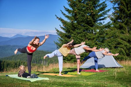 Eine Gruppe von Yoga-Teilnehmern posiert im Freien beim Zelten in den Bergen. Erwachsene und Kinder stehen auf Yogamatten und posieren morgens unter strahlend blauem Himmel.
