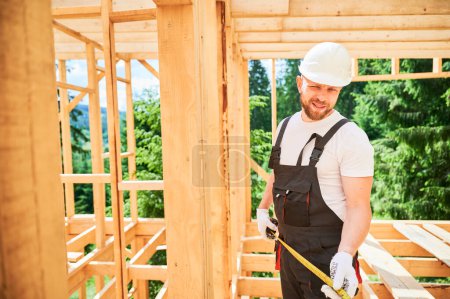 Carpenter constructing wooden-framed house. Portrait of happy man measures distances using tape measure, dressed in work attire and helmet. Concept of ecologically sound, contemporary construction.