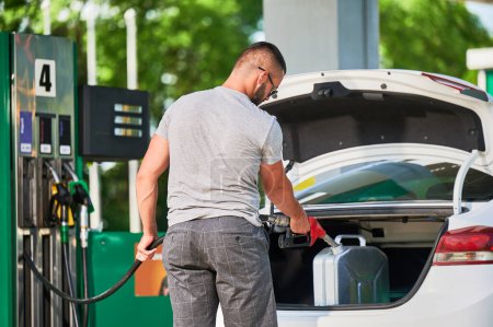 Young man refueling canister with fuel. Skilled driver filling cistern with gasoline in case of unforeseen circumstances. Male adult with pump nozzle refueling canister in car trunk.