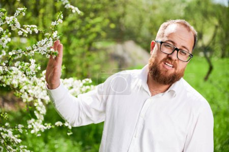 Photo for Man allergic suffering from seasonal allergy at spring in blossoming garden at springtime. Man in glasses and white shirt pushing away branch of blooming tree with flowers. Spring allergy concept - Royalty Free Image