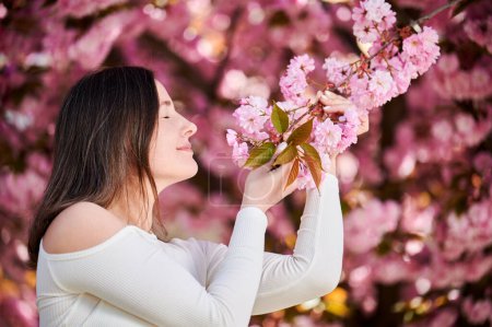 Woman allergic enjoying after treatment from seasonal allergy at spring. Portrait of happy beautiful woman sniffing the flowers in front of blooming sakura tree at springtime. Spring allergy concept.