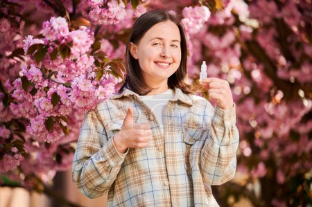 Photo for Woman using medical eyes drops, suffering from seasonal allergy at spring in blossoming garden. Portrait of happy woman showing thumbs up gesture in front of blooming tree. Spring allergy concept. - Royalty Free Image