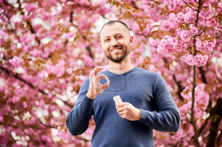 Photo for Man allergic suffering from seasonal allergy in blossoming garden at spring. Handsome smiling man holding allergy pill, making OK hand sign in front of blooming tree. Spring allergy concept. - Royalty Free Image