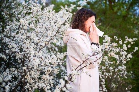 Photo for Man allergic suffering from seasonal allergy at spring in blossoming garden at springtime. Young man sneezing and blowing nose with nasal handkerchief in front of blooming tree. Spring allergy concept - Royalty Free Image