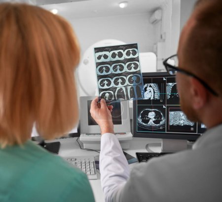 Medical computed tomography or MRI scanner. Back view of doctor and nurse sitting at computer, examining MRI results. Doctor holding scan, showing, studying, examining. Concept of modern diagnostics.