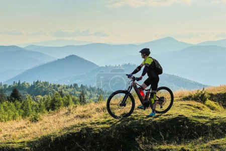 Cyclist man riding electric bike outdoors on sunny day. Male tourist resting on grassy hill, enjoying beautiful mountain landscape, wearing helmet and backpack. Concept of active leisure.