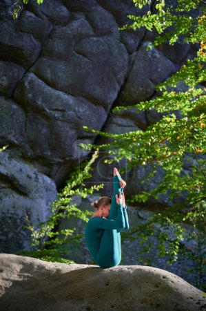 Téléchargez les photos : Femme pratiquant le yoga en plein air dans la forêt. Femme pieds nus sur tapis de yoga entouré d'arbres et de grandes roches, ce qui suggère un environnement paisible et naturel idéal pour la méditation ou la pratique du yoga. - en image libre de droit