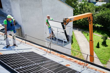 Workers lifting up photovoltaic solar panel on metal rooftop of house with assistance of crane lift. Men installers installing photovoltaic solar modules outdoors. Renewable energy generation concept.