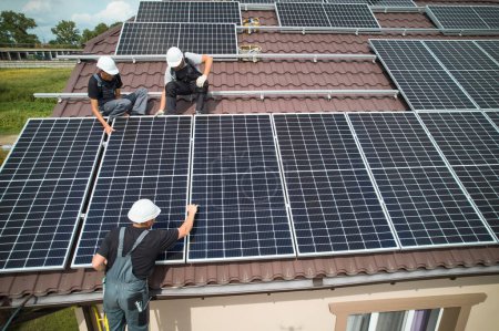 Téléchargez les photos : Hommes travailleurs installant des modules solaires photovoltaïques sur le toit de la maison. Ingénieurs dans la construction de casques système de panneaux solaires à l'extérieur. Concept d'énergies alternatives et renouvelables. Vue aérienne. - en image libre de droit