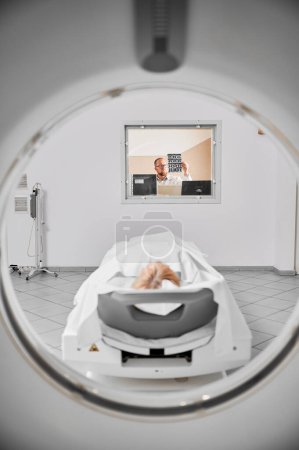 Medical computed tomography or MRI scanner. Doctor examining MRI results, female patient lying on couch. Doctor wearing glasses, holding MRI results. Concept of modern diagnostics.