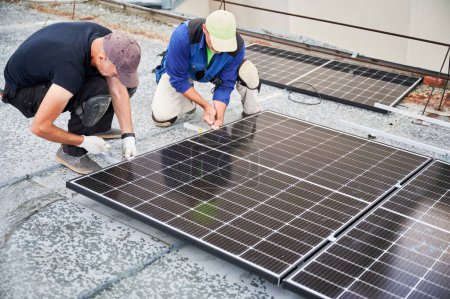 Téléchargez les photos : Les travailleurs construisent un système de panneaux solaires photovoltaïques sur le toit métallique de la maison. Des ingénieurs hommes installent un module solaire à l'aide d'une clé à molette à l'extérieur. Concept de production d'énergies alternatives et renouvelables. - en image libre de droit