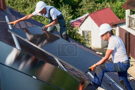 Téléchargez les photos : Les travailleurs construisent un système de panneaux solaires sur le toit de la maison. Deux hommes installateurs de casques installant le module solaire photovoltaïque à l'extérieur. Concept de production d'énergies alternatives, vertes et renouvelables. - en image libre de droit