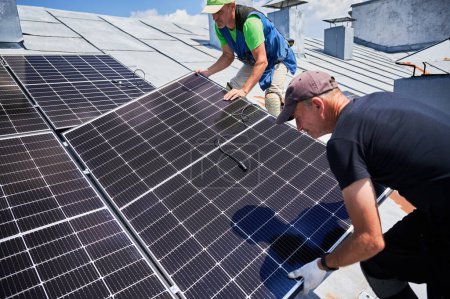 Téléchargez les photos : Les travailleurs construisent un système de panneaux solaires sur le toit métallique de la maison. Deux installateurs hommes installant le module solaire photovoltaïque à l'extérieur. Concept de production d'énergies alternatives, vertes et renouvelables. - en image libre de droit