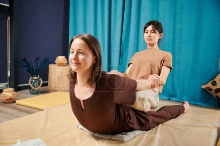 Téléchargez les photos : Masseuse faisant un massage de yoga thaï. Femme thérapeute faisant un traitement de massage traditionnel, étirant le corps des patientes. Client couché sur le tapis, profitant d'un massage des mains dans une atmosphère calme. - en image libre de droit