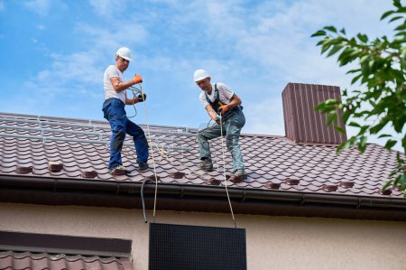 Téléchargez les photos : Hommes travailleurs installant le système de panneaux solaires sur le toit de la maison. Electriciens dans les casques soulevant le module solaire photovoltaïque à l'aide de cordes à l'extérieur. Concept d'énergies alternatives et renouvelables. - en image libre de droit