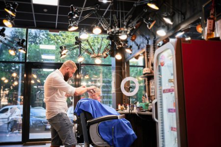 Téléchargez les photos : Homme coiffeur couper les cheveux des petits garçons en utilisant peigne et ciseaux. Un enfant qui se fait couper les cheveux par un homme adulte, probablement un coiffeur. Coiffeur professionnel et client mignon au salon de coiffure moderne. - en image libre de droit