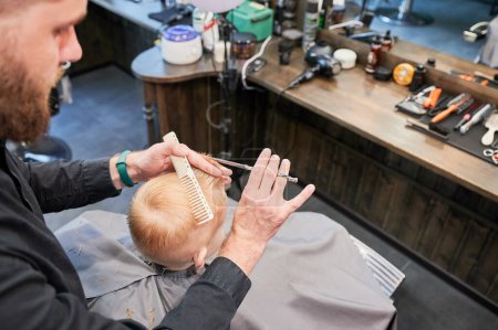Téléchargez les photos : Homme coiffeur couper les cheveux des petits garçons en utilisant peigne et ciseaux. Un enfant qui se fait couper les cheveux par un homme adulte, probablement un coiffeur. Coiffeur professionnel et client mignon au salon de coiffure moderne. - en image libre de droit
