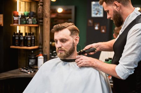 Téléchargez les photos : Bearded handsome client getting stylish haircut. Professional barber shaving clients nape. Confident man on regular appointment in barbershop. - en image libre de droit