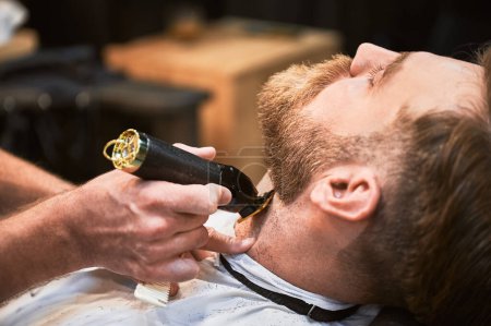 Téléchargez les photos : Close-up of professional barber trimming clients beard. Male hairdresser serving client with thin beard by clipper. Isolated photo of man getting his haircut done. - en image libre de droit