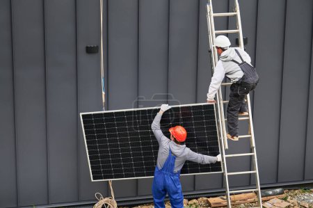 Männer Bauarbeiter installieren Solaranlage auf dem Dach des Hauses. Elektriker in Helmen, die mit Hilfe von Seilen im Freien Photovoltaik-Solarmodule hochziehen. Konzept der alternativen und erneuerbaren Energien.