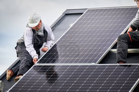 Téléchargez les photos : Electriciens construisant la station de module solaire photovoltaïque sur le toit de la maison. Hommes techniciens dans les casques installant le système de panneaux solaires à l'extérieur. Concept d'énergies alternatives et renouvelables. - en image libre de droit