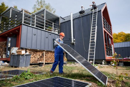 Téléchargez les photos : Roofers installing solar panel system on roof of house. Man worker in helmet carrying photovoltaic solar module outdoors. Concept of alternative and renewable energy. - en image libre de droit