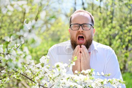 Téléchargez les photos : Homme allergique souffrant d'allergie saisonnière au printemps dans le jardin en fleurs au printemps. Jeune homme barbu avec des lunettes éternuant devant un arbre en fleurs. Concept d'allergie printanière - en image libre de droit