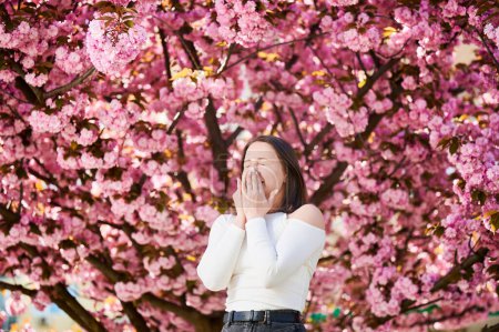 Photo for Woman allergic suffering from seasonal allergy at spring in blossoming garden at springtime. Young woman sneezing, having runny nose in front of blooming tree. Spring allergy concept - Royalty Free Image