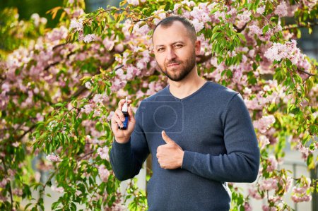 Homme allergique à l'aide de gouttes nasales médicales, souffrant d'allergie saisonnière au printemps dans le jardin en fleurs. Un bel homme souriant montrant des pouces près d'un arbre en fleurs à l'extérieur. Concept d'allergie printanière.