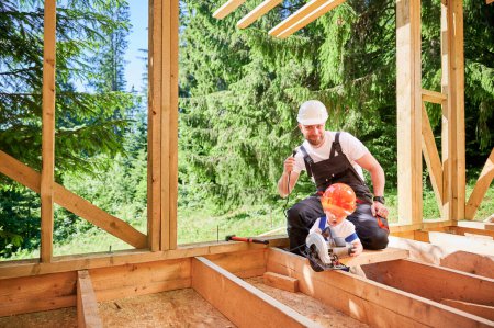 Téléchargez les photos : Father with toddler son building wooden frame house. Man playing with his son using electric saw, demonstrating that it unplugged from power source on construction site. Carpentry and family concept. - en image libre de droit