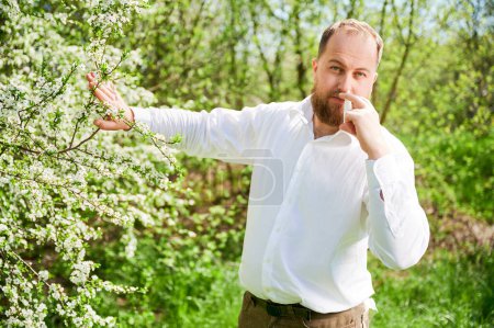 Téléchargez les photos : Man allergic using medical nasal spray, suffering from seasonal allergy at spring in blossoming garden. Handsome man treating runny nose in front of blooming tree outdoors. Spring allergy concept. - en image libre de droit