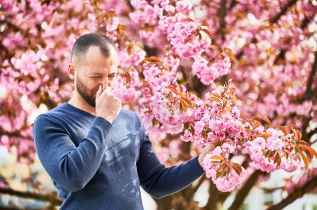 Photo for Man allergic suffering from seasonal allergy at spring in blossoming garden at springtime. Bearded young man sneezing and having runny nose in front of blooming tree. Spring allergy concept. - Royalty Free Image