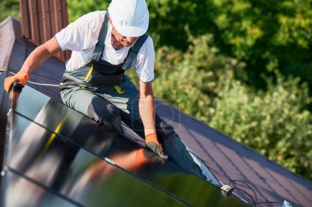 Téléchargez les photos : Travailleur construction système de panneaux solaires sur le toit de la maison. Homme ingénieur dans le casque installant le module solaire photovoltaïque à l'extérieur. Concept de production d'énergies alternatives, vertes et renouvelables. - en image libre de droit