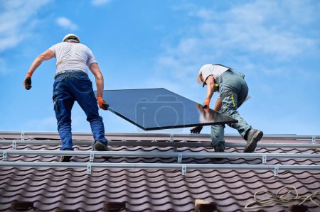 Téléchargez les photos : Les travailleurs construisent un système de panneaux solaires sur le toit de la maison. Deux hommes installateurs dans des casques transportant module solaire photovoltaïque à l'extérieur. Concept de production d'énergies alternatives, vertes et renouvelables. - en image libre de droit