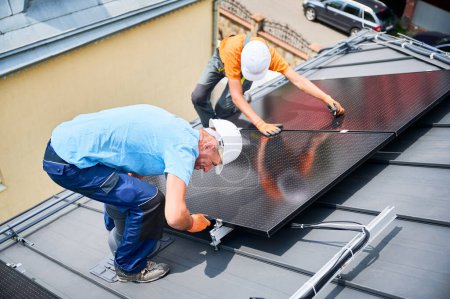 Téléchargez les photos : Les travailleurs construisent un système de panneaux solaires photovoltaïques sur le toit de la maison. Hommes techniciens en casques et gants installant le module solaire avec l'aide de clé hexagonale à l'extérieur. Concept de production d'énergie renouvelable. - en image libre de droit