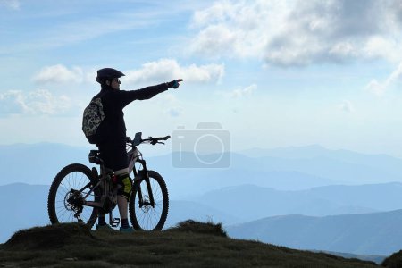 Cyclist man riding electric bike outdoors. Silhouette of male tourist resting on top of hill, pointing on beautiful mountain landscape, wearing helmet and backpack. Concept of active leisure.