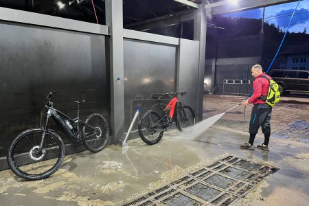 Cyclist using high-pressure water spray to clean two electric mountain bikes on outdoor washing station.