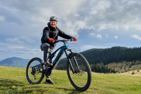 Cyclist man riding electric bike outdoors. Portrait of smiling male tourist resting on grassy hill, enjoying beautiful mountain landscape, wearing helmet. Concept of active leisure.