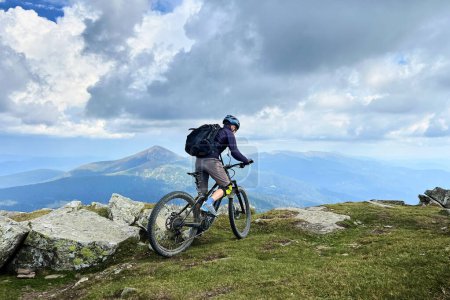 Cyclist man riding electric mountain bike outdoors. Male tourist biking along grassy trail in the mountains, wearing helmet and backpack. Concept of sport, active leisure and nature.
