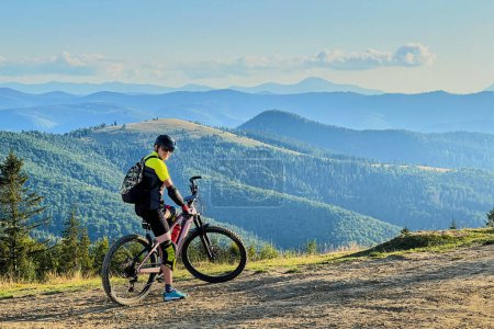 Cyclist man riding electric bike outdoors on sunny day. Male tourist resting on hill, enjoying beautiful mountain landscape, wearing helmet and backpack. Concept of active leisure.