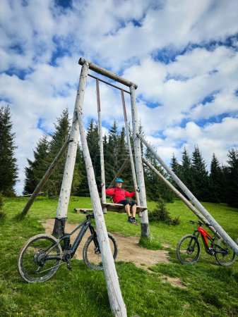 Persona sentada en una gran estructura oscilante de madera en una colina o montaña, adecuada para actividades recreativas al aire libre. Dos bicicletas eléctricas de montaña apoyadas en el bastidor basculante.