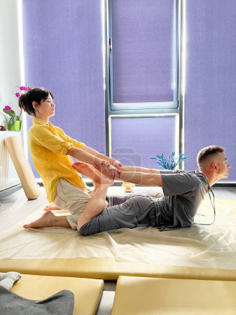 Masajista haciendo masaje tailandés de yoga. Mujer terapeuta haciendo el tratamiento de masaje tradicional, estirando el cuerpo de los pacientes masculinos. Cliente acostado en la alfombra, disfrutando del masaje de manos en un ambiente tranquilo.