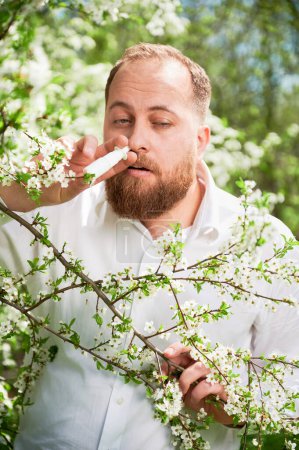Man allergic using medical nasal drops, suffering from seasonal allergy at spring in blossoming garden. Handsome man treating runny nose in front of blooming tree outdoors. Spring allergy concept.
