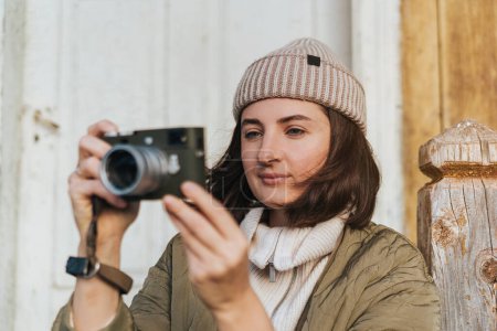 Photo for Happy smiling girl wearing beanie hat taking pictures by vintage photo camera. Portrait of young travel woman sitting on the porch of an old house. - Royalty Free Image