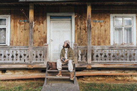 Téléchargez les photos : Portrait de jeune voyageuse relaxante assise sur le porche d'une vieille maison et profitant de la vie dans les montagnes. - en image libre de droit