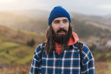 Photo for Portrait of hiker - man hiking on mountains. Handsome male traveler photographer looking to the side walking on hills wearing blue beanie, plaid shirt, orange sweatshirt and with a leather backpack on shoulders. - Royalty Free Image