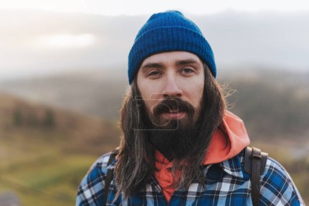 Photo for Handsome male traveler photographer looking to the side walking on hills wearing blue beanie, plaid shirt, orange sweatshirt and with a leather backpack on shoulders. Portrait of hiker - man hiking on mountains. - Royalty Free Image